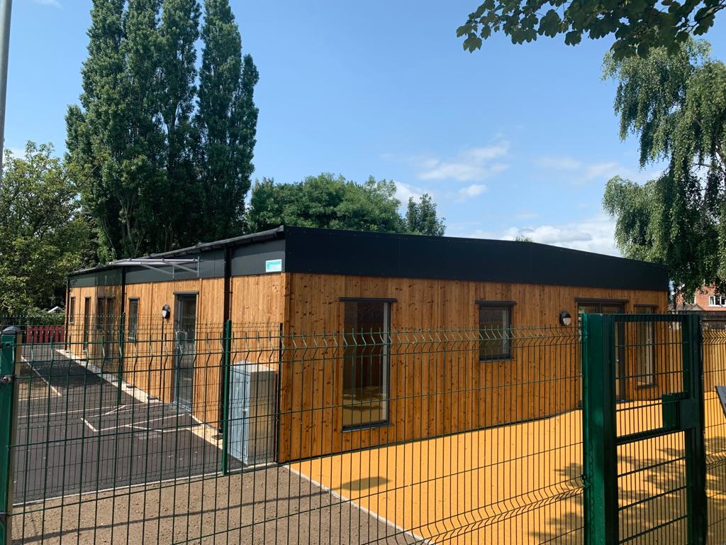 Large modular building with external wooden cladding. The building has large full length windows and is sat behind a fence.