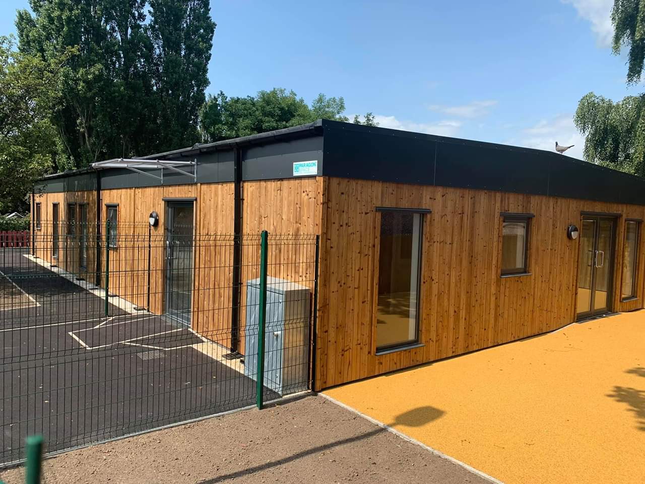 Large modular building with external wooden cladding. The building has large full length windows and is sat behind a fence.